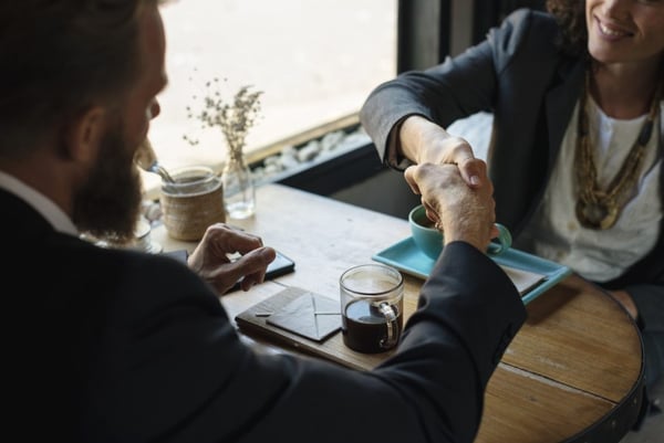 man and woman shake hands over a table with coffee mugs in front of them